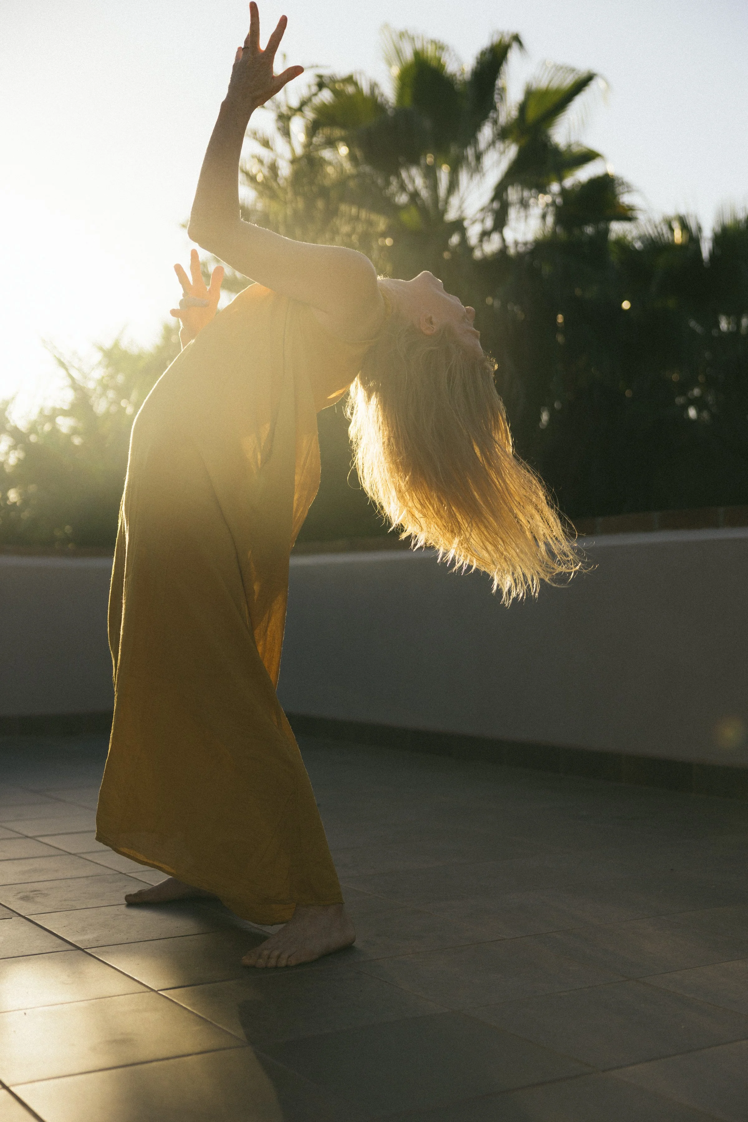A person dressed in a long yellow dress practicing dance or yoga outdoors during sunset, with palm trees in the background.