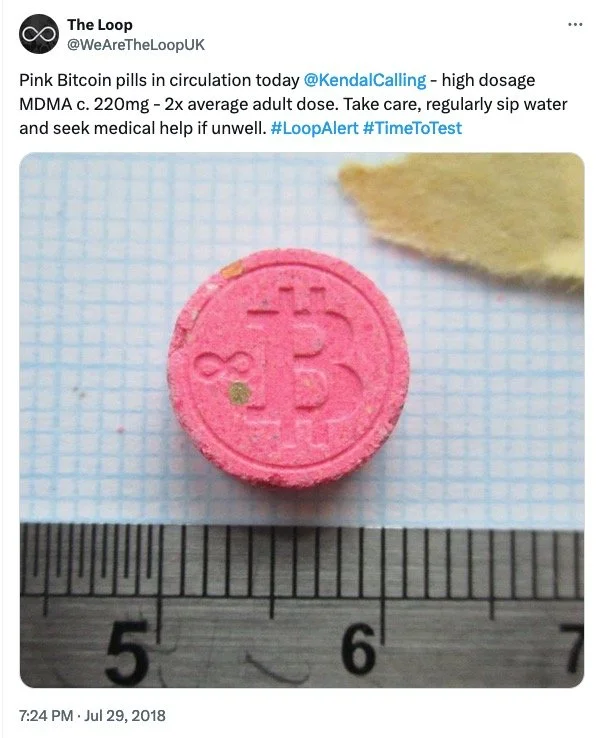 A pink Bitcoin pill tablet with the Bitcoin symbol embossed on it, placed on graph paper with a ruler for scale, near a piece of yellowish paper.