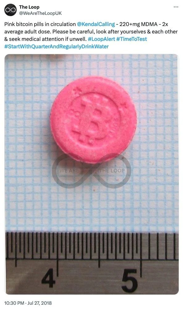 Pink pill with a Bitcoin symbol in the center, placed on graph paper with a ruler below for scale.