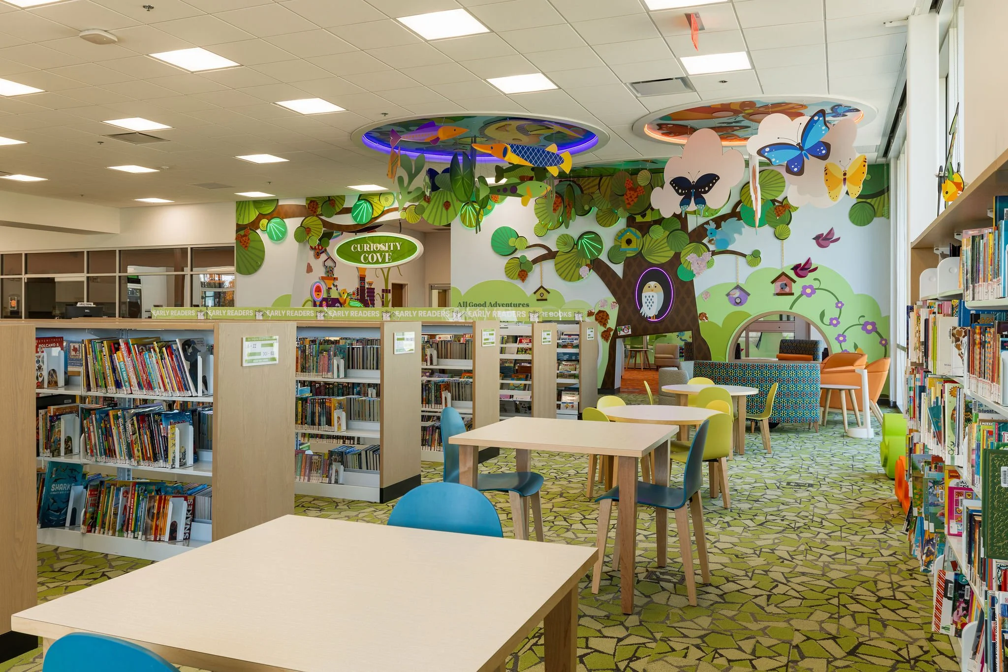 Children's section of a library with colorful wall murals featuring trees, butterflies, birds, and frogs, and bookshelves filled with books.