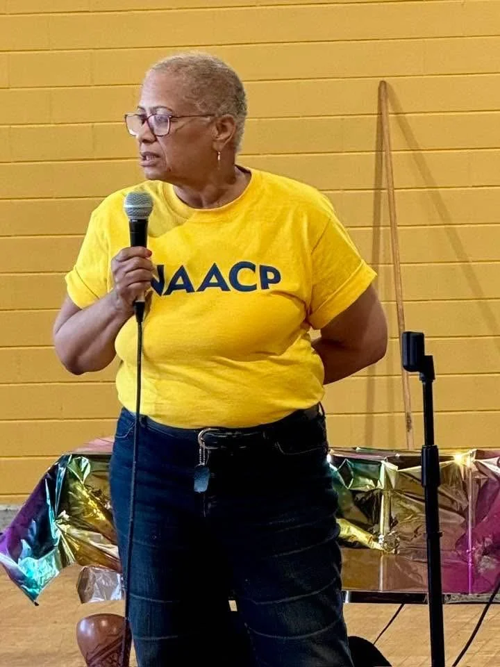 An older woman wearing glasses and a yellow NAACP t-shirt holding a microphone at a public event, with a yellow wall and colorful decorations in the background.