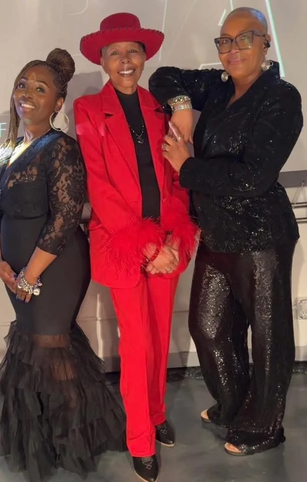 Three women wearing formal attire, standing close together and smiling at an indoor event.