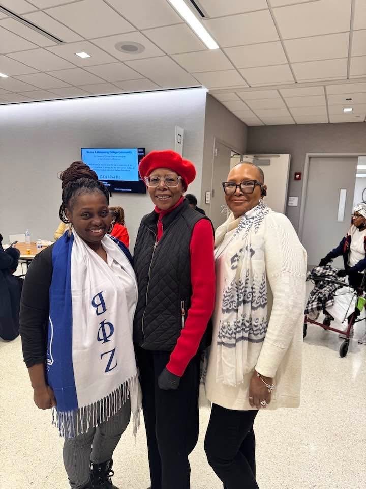 Three women standing together indoors, smiling at the camera. The woman on the left is wearing a black cardigan, gray pants, and a white scarf with blue Greek letters. The woman in the middle is wearing a red beret, glasses, a black quilted vest over
