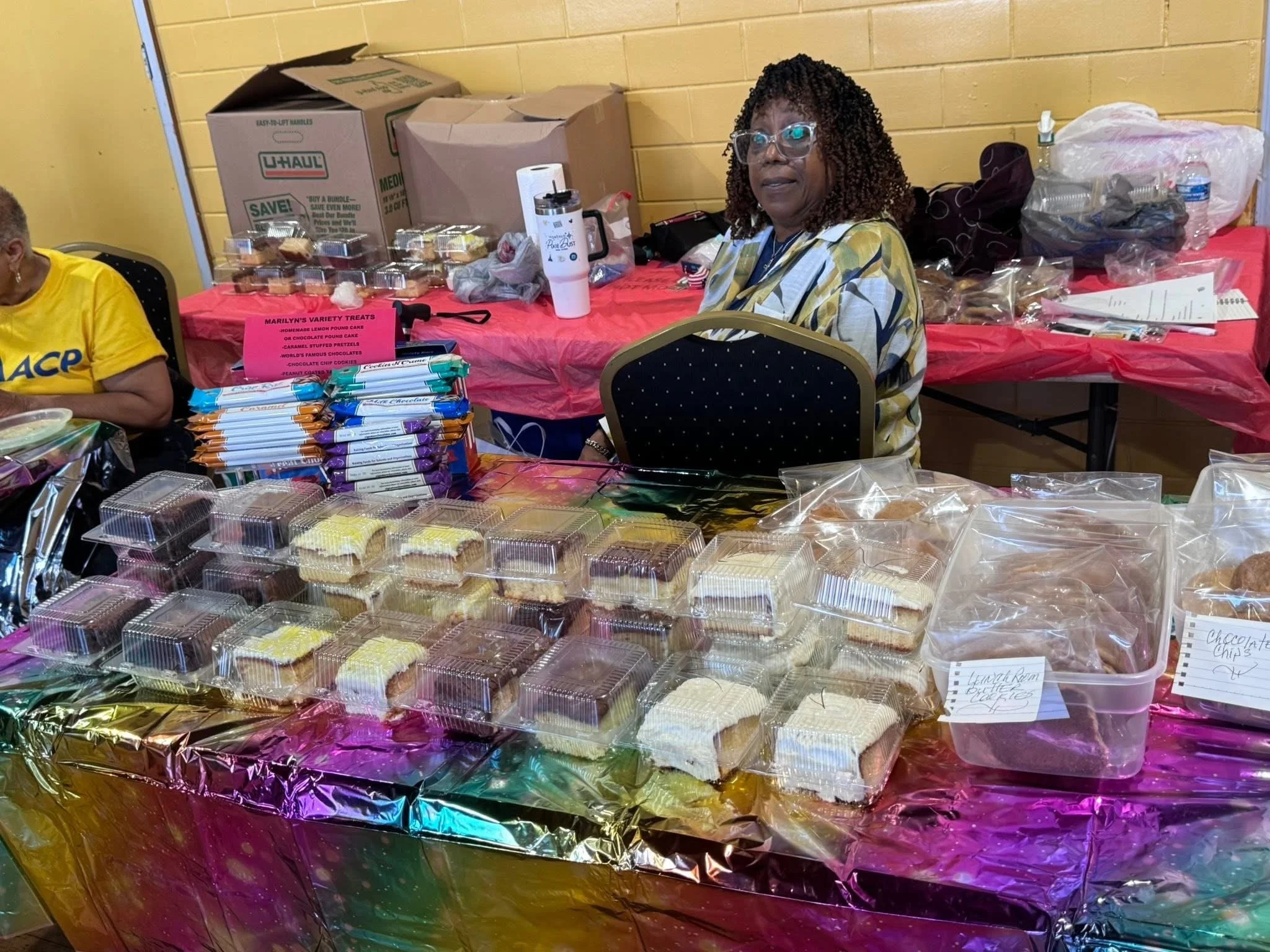 A woman with curly hair and glasses sitting at a table covered with a colorful tablecloth, surrounded by baked goods such as cupcakes, cookies, and slices of cake, at an indoor event.
