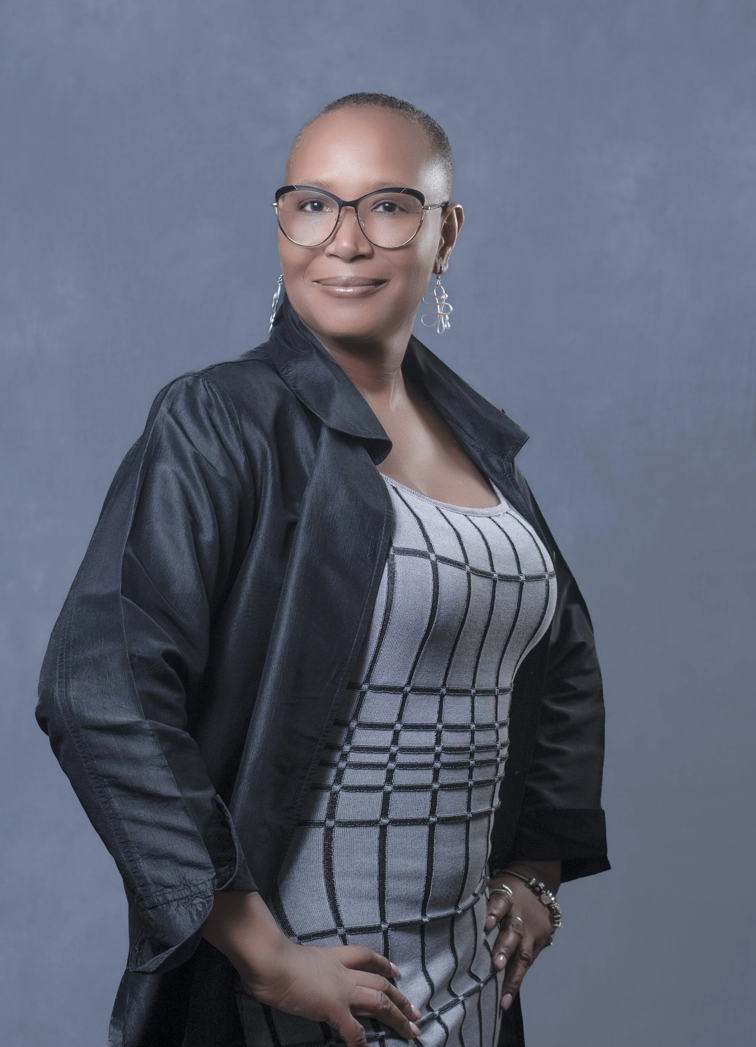 A woman with short hair wearing glasses, earrings, a black satin jacket, and a dress with a plaid pattern, standing against a gray background.