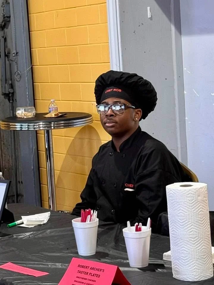 A young person dressed as a chef, wearing glasses, a black chef's uniform, and a chef's hat with 'Chef Rob' embroidered on it, sitting at a table with condiments and paper products in a room with yellow brick walls.