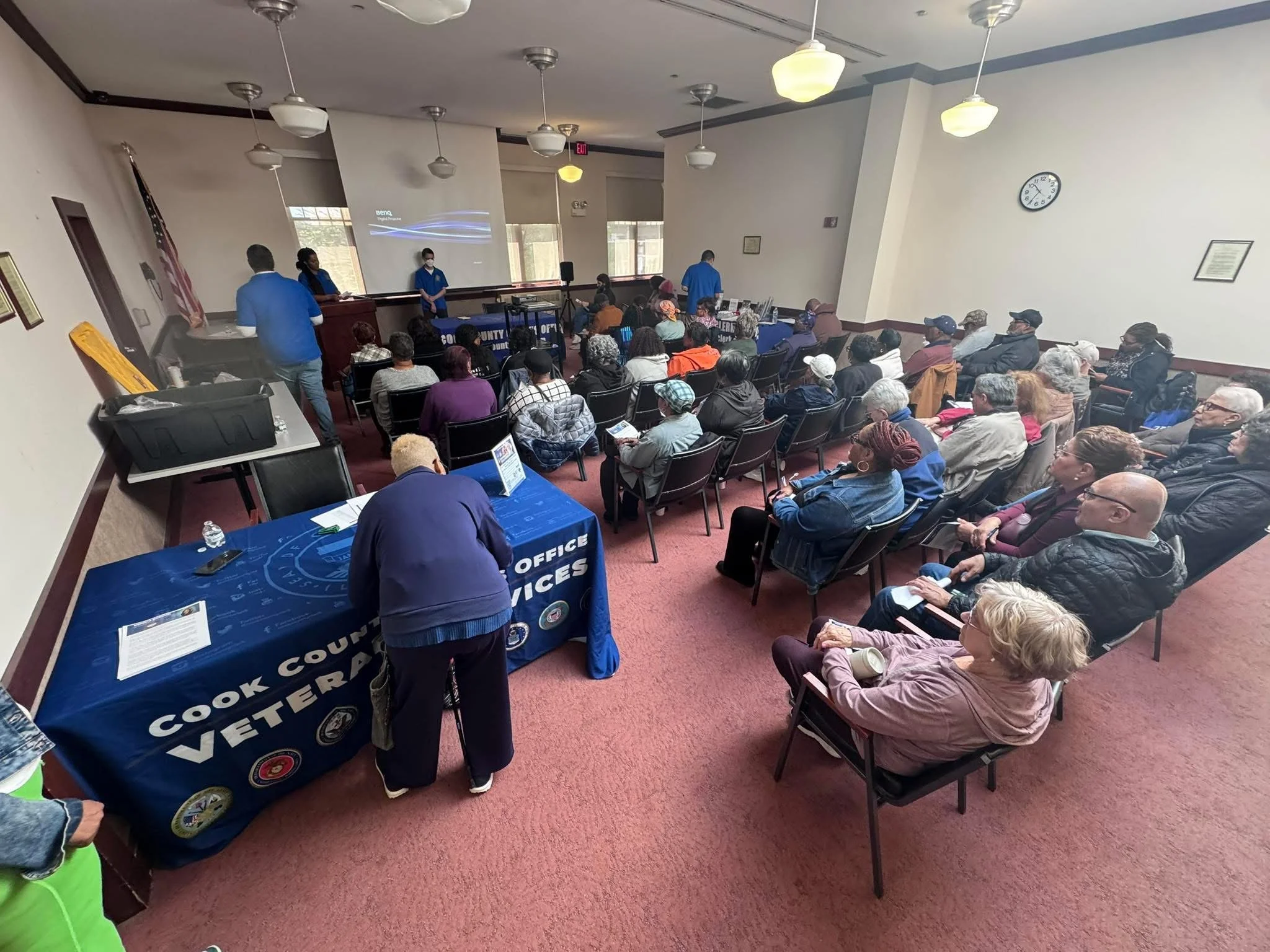 Scene of a community event or town hall meeting with a diverse group of attendees seated and listening to presenters. There are tables with informational materials and a banner that reads 'Cook County Office of the Voter' indicating a voting or elect