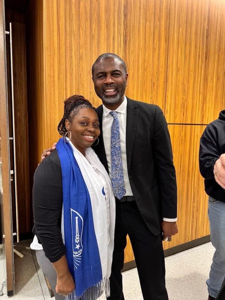 A woman and a man smiling and standing close together, with the woman wearing a blue and white scarf and the man dressed in a dark suit and tie, in front of wooden paneling.