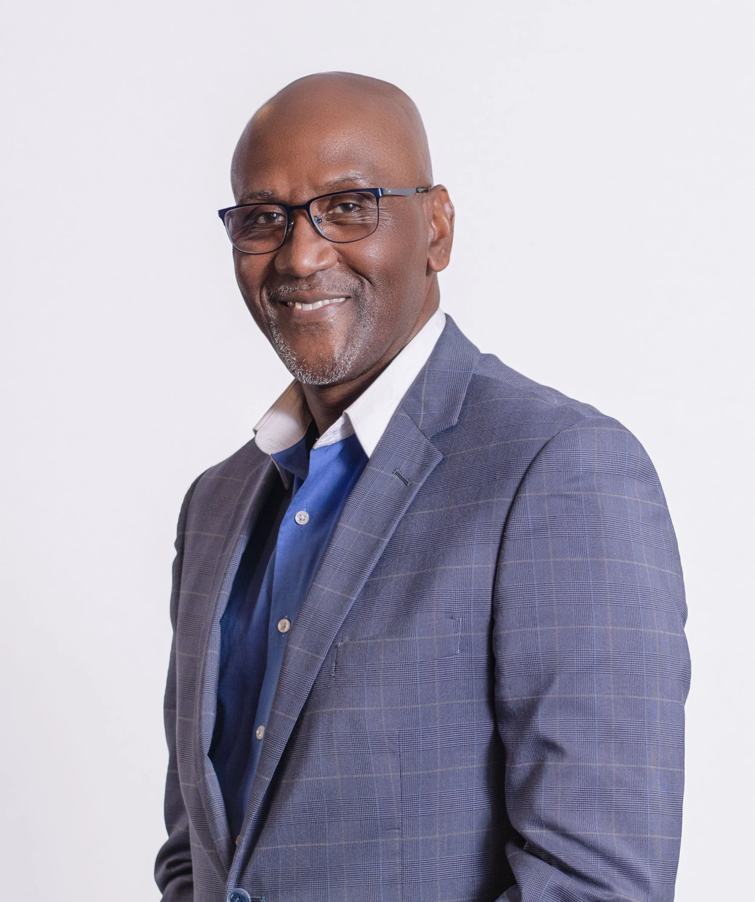 A middle-aged Black man wearing glasses, a white shirt, and a blue plaid blazer, smiling against a plain white background.