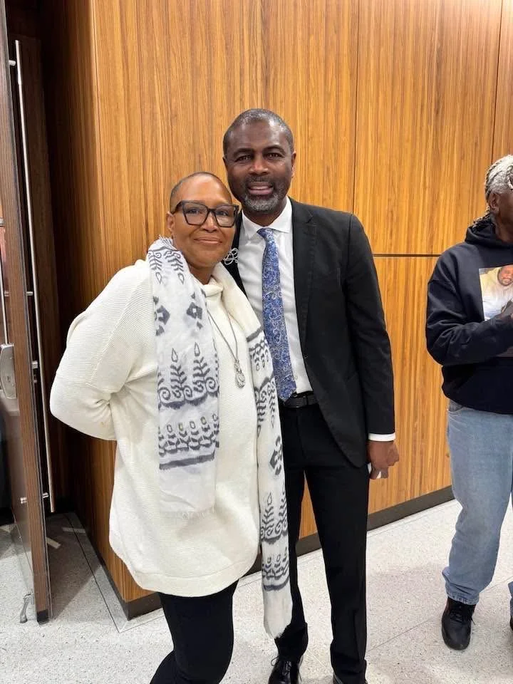 A woman and a man standing together, smiling indoors near a wooden wall. The woman wears glasses, a white scarf with black patterns, and a white top. The man wears a black suit, white shirt, and a patterned blue tie.