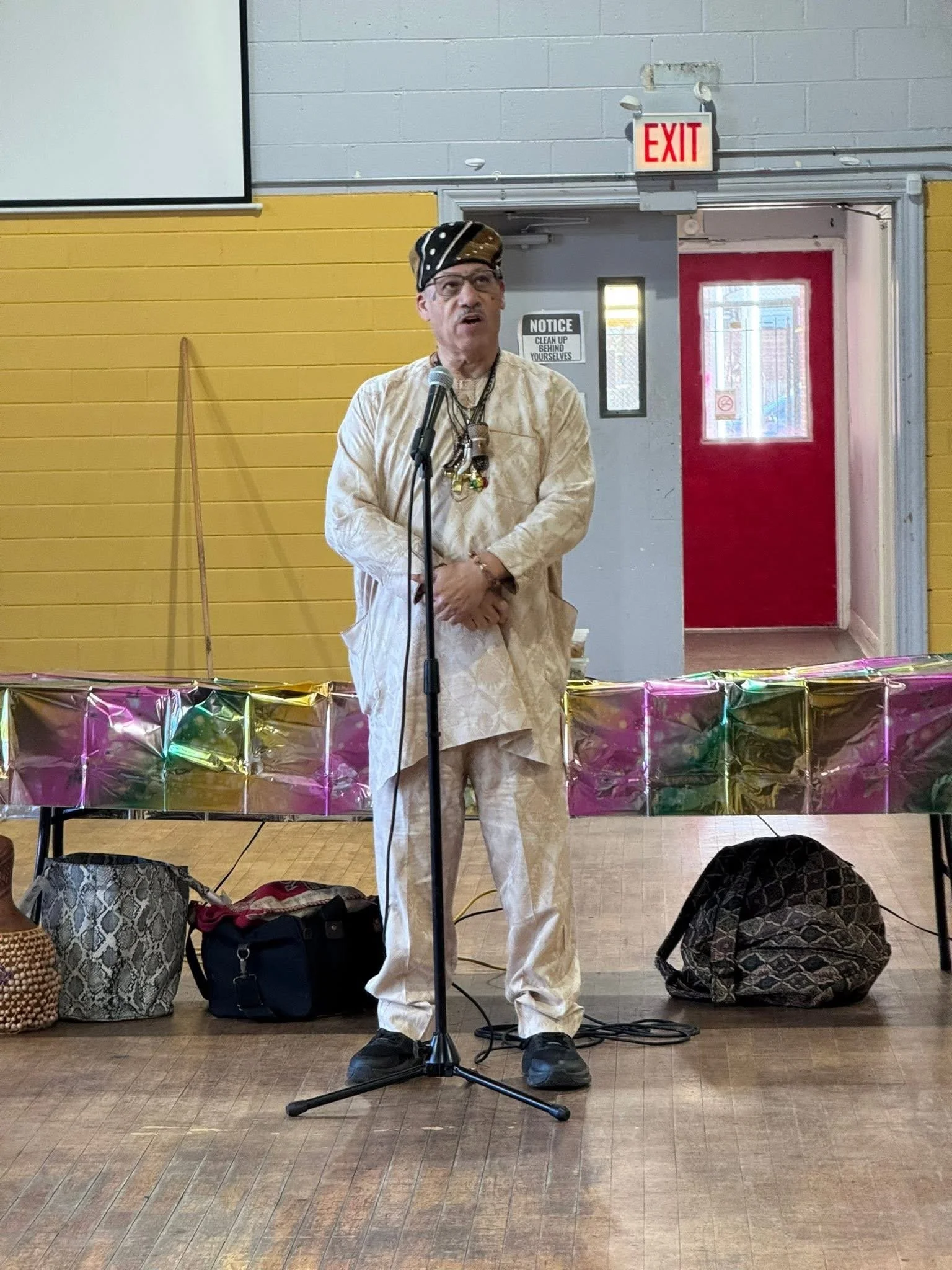 A man speaking into a microphone on stage, dressed in traditional attire, with bags on the floor and colorful decorations in the background.