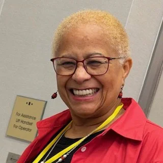 An elderly woman with short blonde hair and glasses, smiling, wearing a red blazer and yellow top, standing indoors.