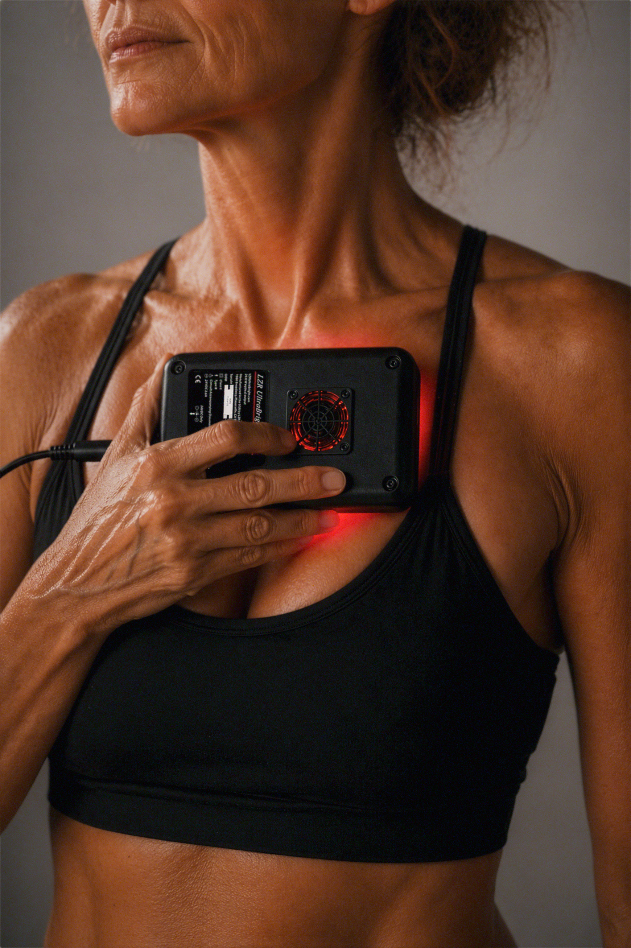 Close-up of a woman with dark skin in athletic attire, holding a device against her chest emitting red light, with focus on her neck and shoulders.