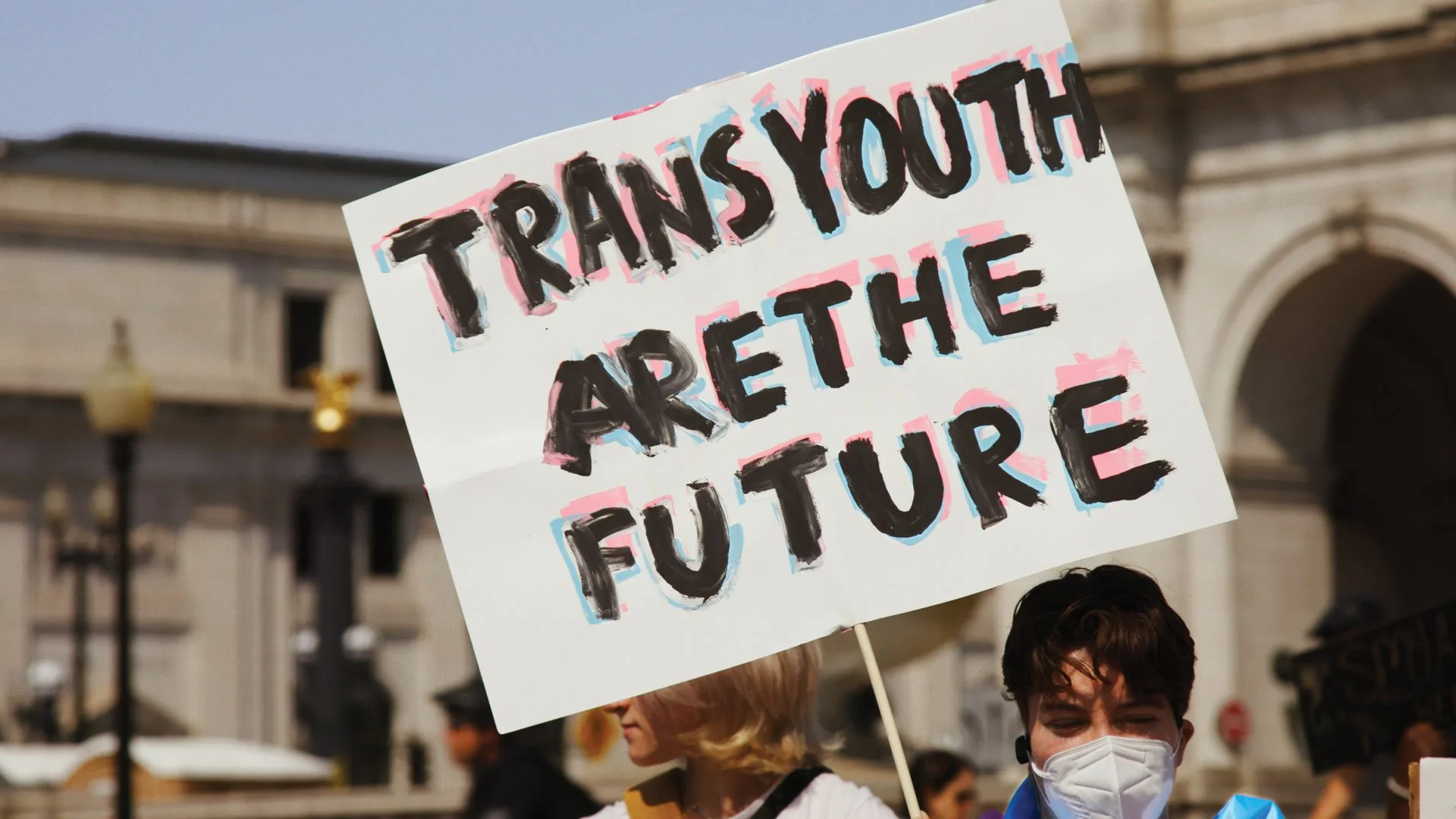 Person holding a protest sign that reads 'Trans Youth Are The Future' with people and buildings in the background.