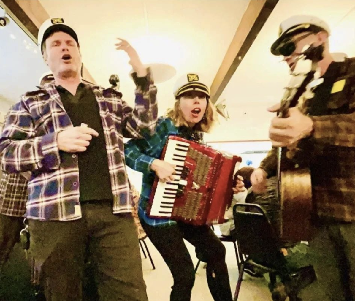 Three people in a room singing sea shanties and playing musical instruments, wearing captain's hats, with festive lighting in the background.