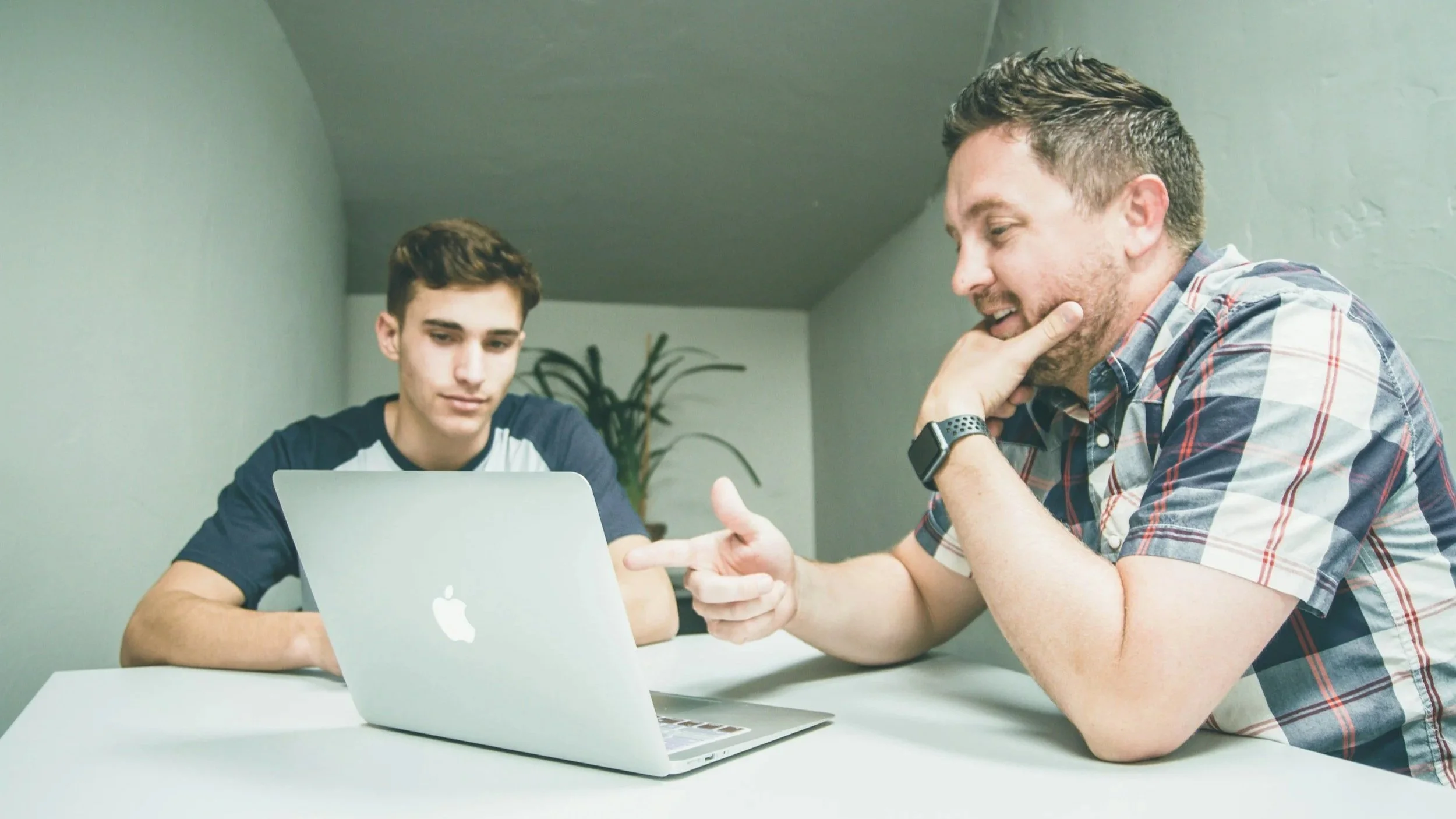 Two men sitting at a white table, working on a silver MacBook laptop, having a conversation, with a green wall and plant in the background.