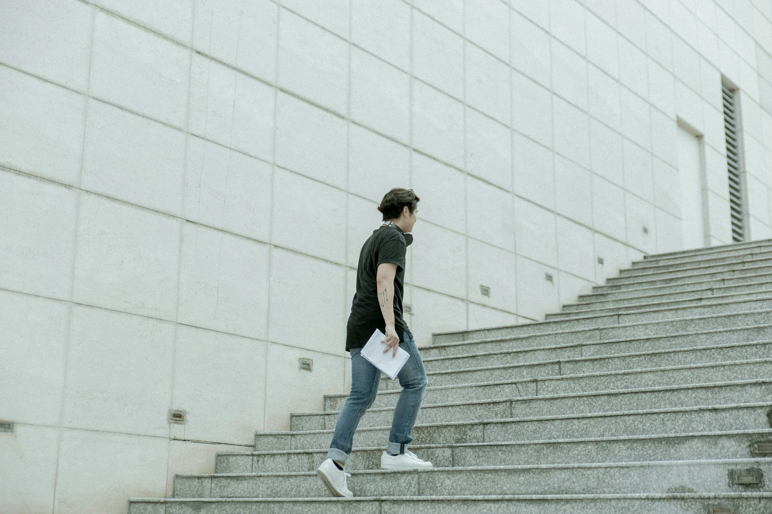 A person with short dark hair, wearing a black t-shirt, jeans, and white shoes, walking up concrete stairs while holding a white paper or notebook in their right hand, in front of a large light-colored brick wall with steps and a ventilation grille.