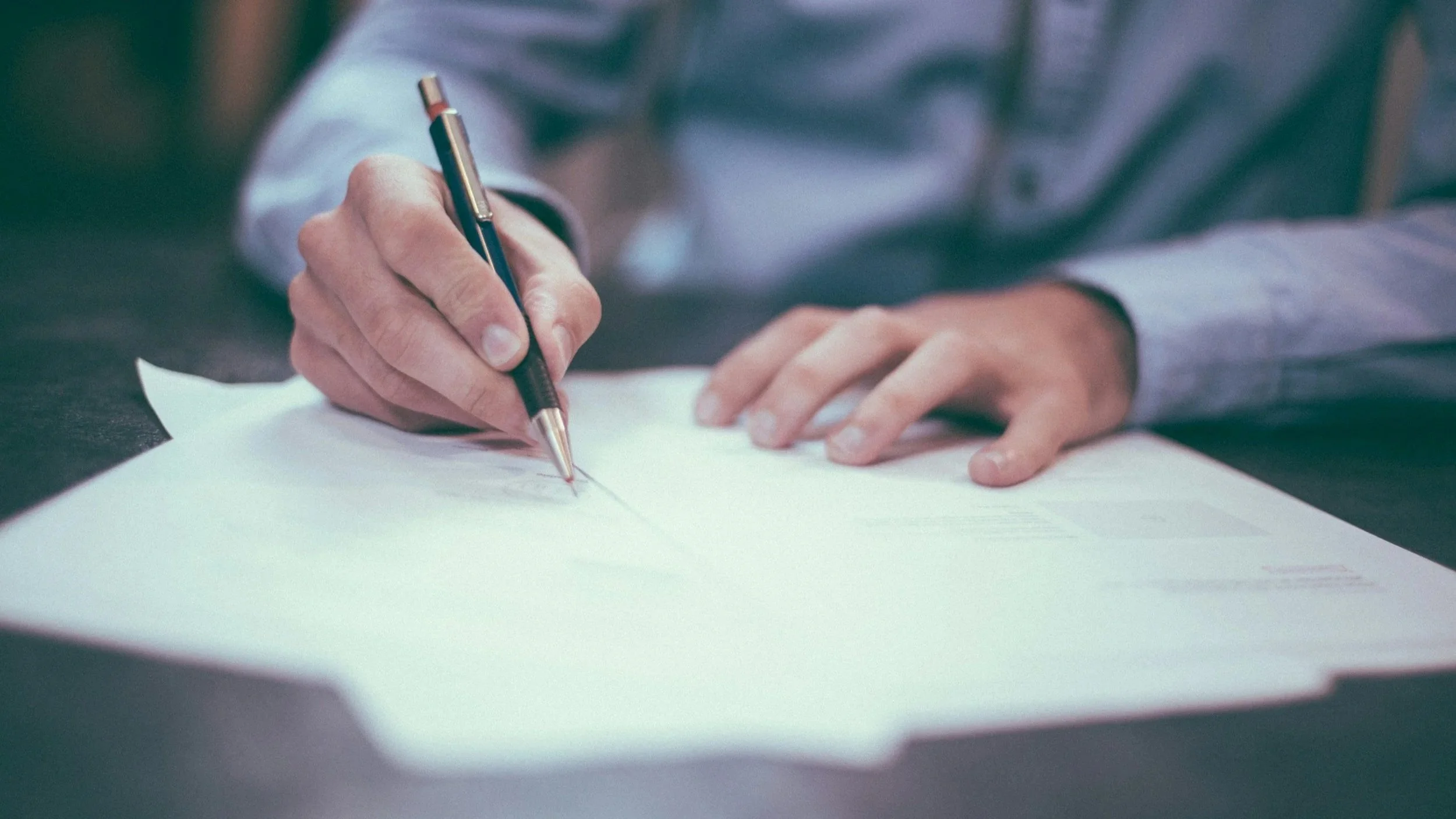 A person in a light blue shirt is writing on a piece of paper with a black and silver pen, resting on a dark surface.