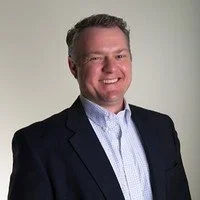 A smiling man with short hair in a dark blazer and light shirt against a plain background.