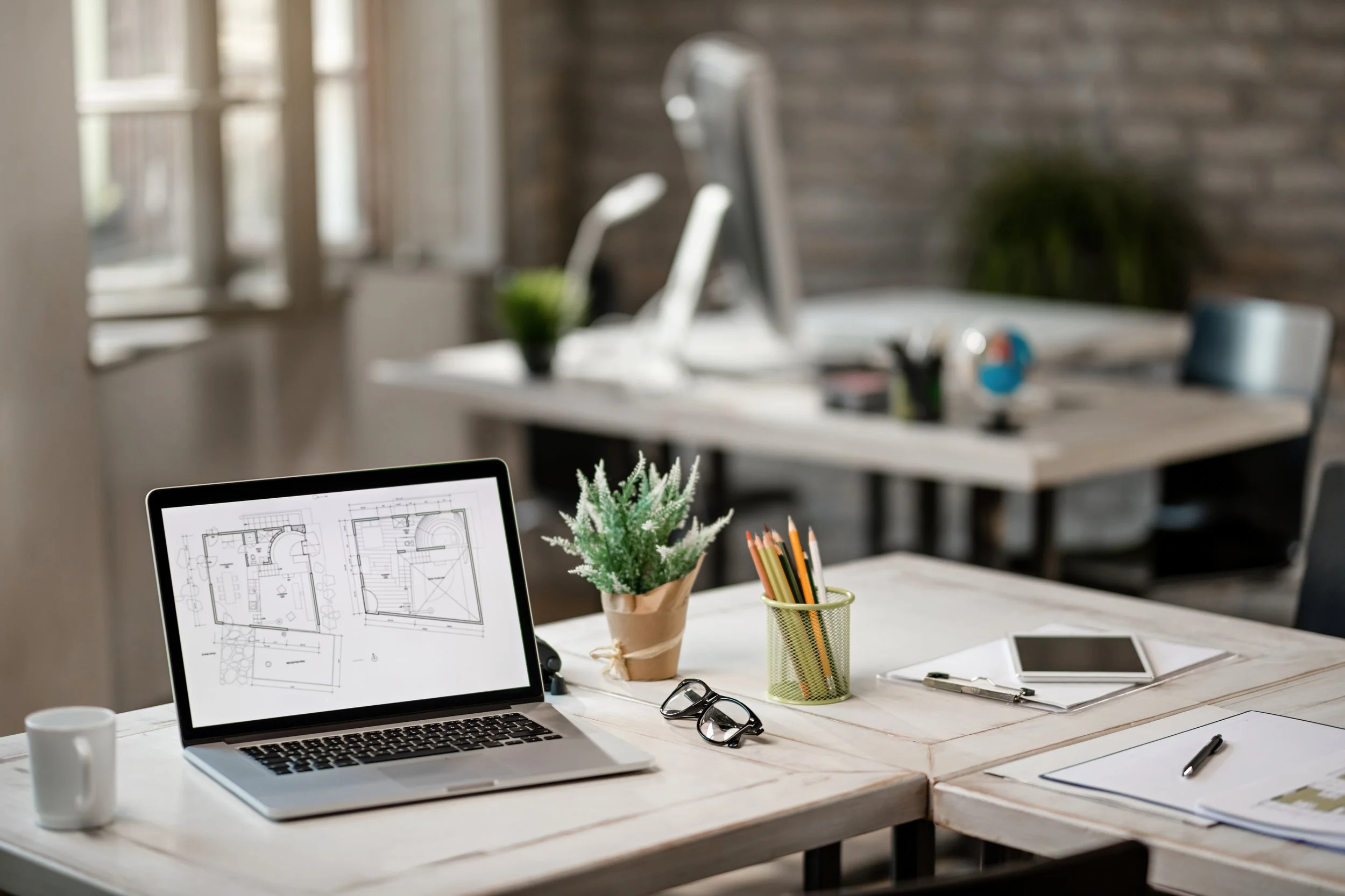 Open laptop displaying a floor plan on a white wooden table, with a coffee mug, glasses, a potted plant, and a cup holding pencils and pens. In the background, there is an office space with a white desk, a computer monitor, and some chairs, with a brick wall and a window.