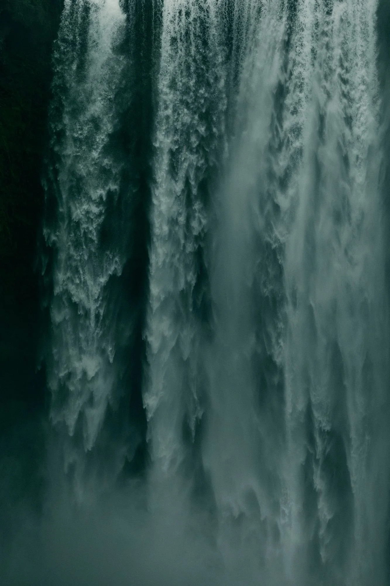 A dramatic, vertical close-up photograph of a rushing green-toned waterfall cascading down with heavy mist and foam.