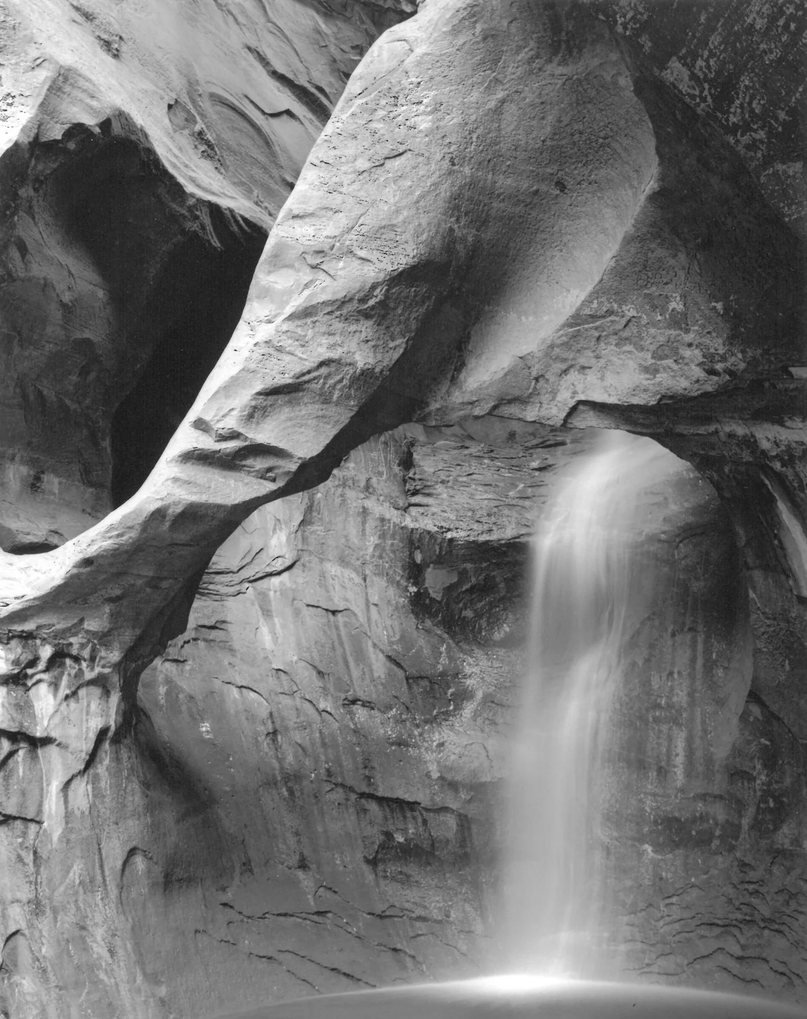 A dramatic black and white photograph of a hidden grotto waterfall cascading through a massive natural stone archway into a quiet pool, highlighting the deep textures of the surrounding canyon rock walls.