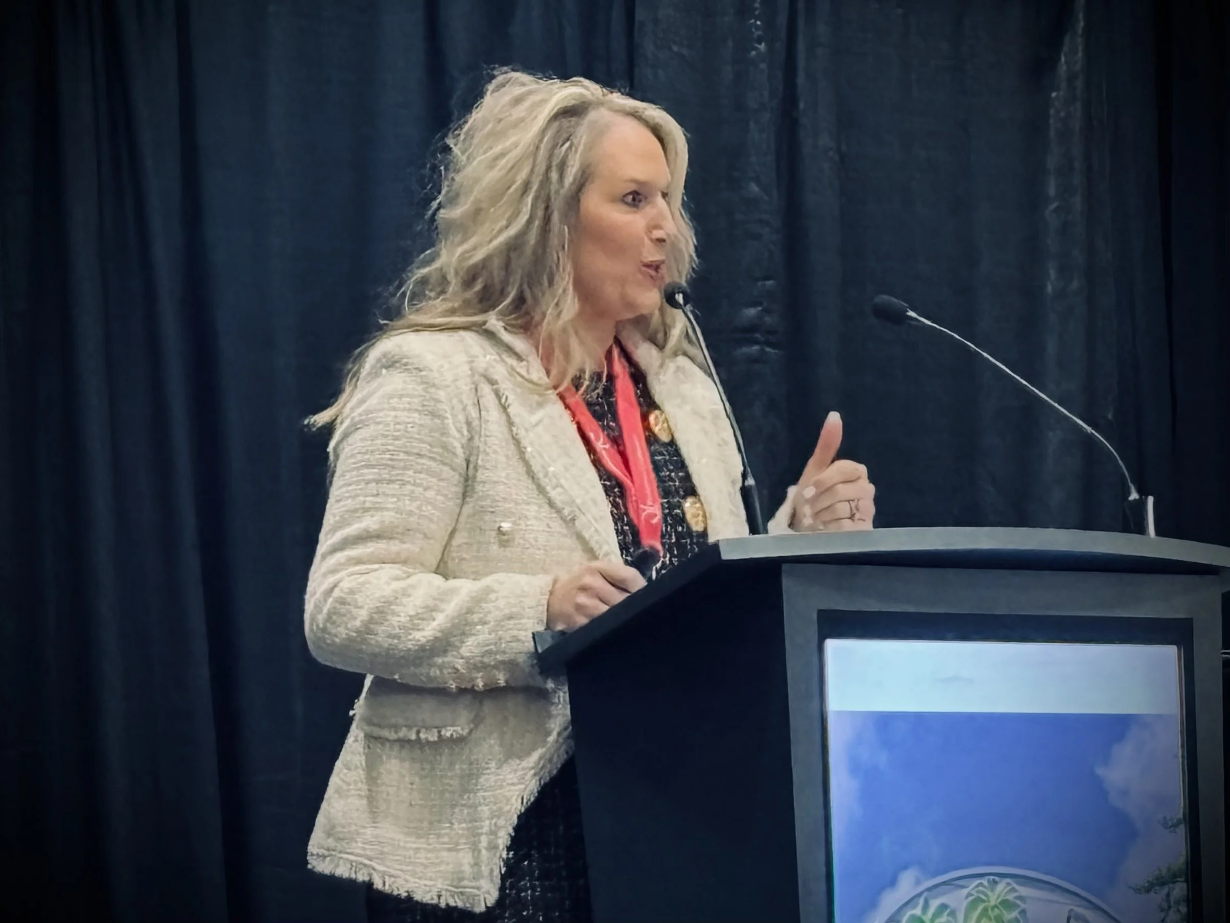 A woman with long blonde hair speaking at a podium during a conference, with black curtains in the background.