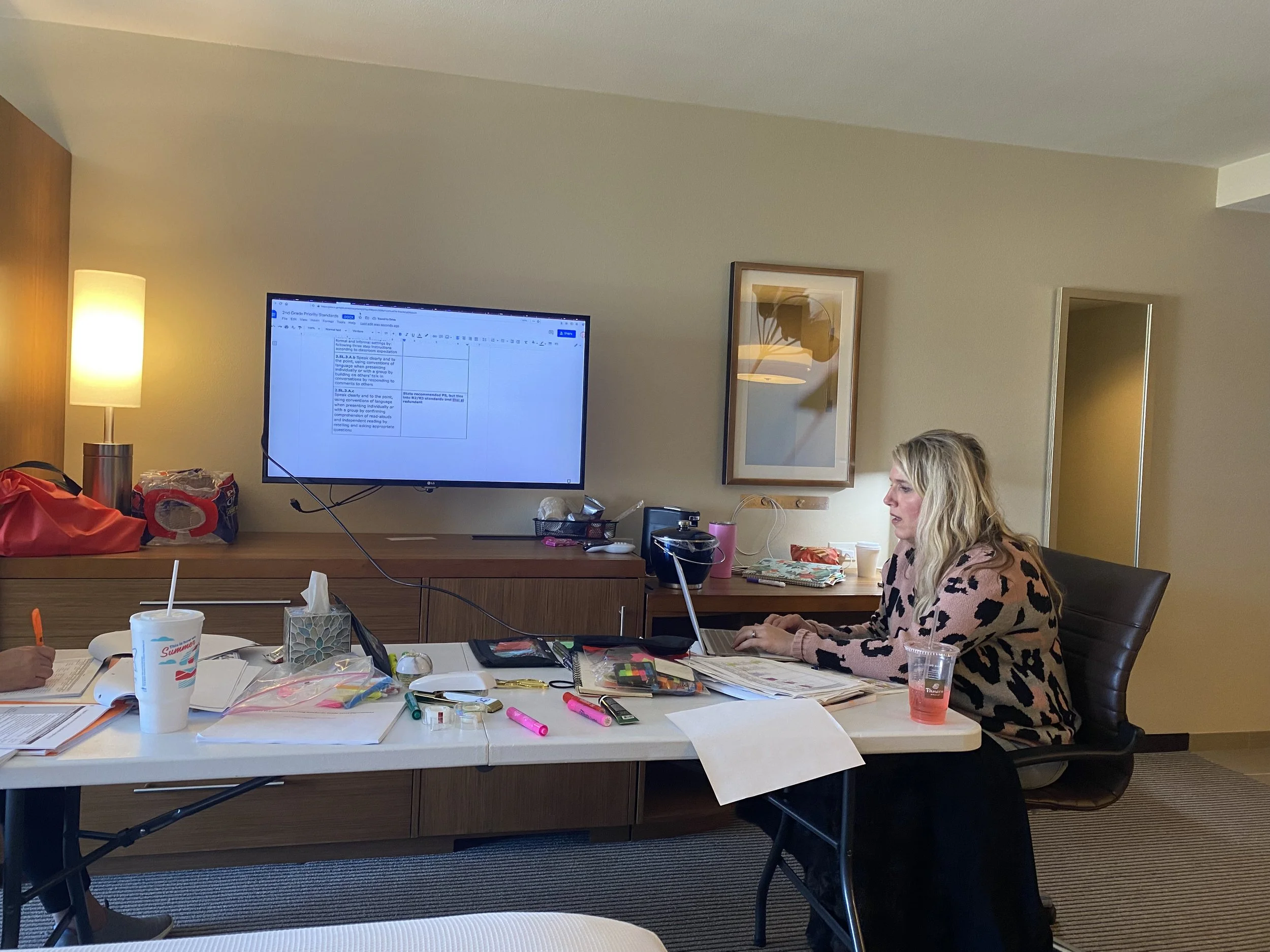 A woman with blonde curly hair working on a laptop at a loosely organized conference table filled with papers, markers, and drinks. Behind her, a wall-mounted flat-screen monitor displays a document, and a framed piece of artwork hangs on the wall.
