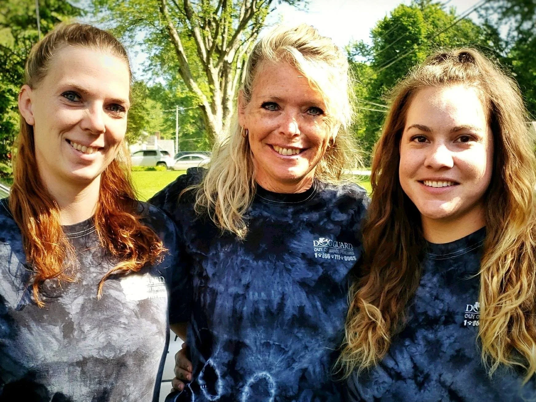 Three women outdoors in a park, smiling, with trees and cars in the background. All wear matching dark tie-dye shirts that say 'Dog Guard' and feature a phone number.