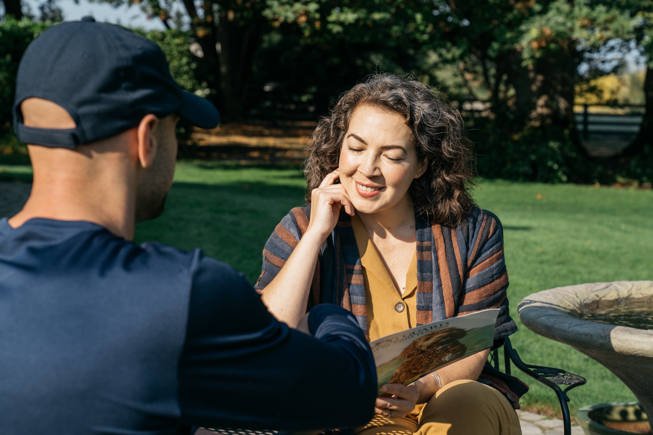 A woman with curly brown hair smiling with closed eyes, sitting outdoors at a table, with her hand gently touching her face, while a man in a navy baseball cap and shirt shows her something in a book or magazine. The background features trees and a grassy area on a sunny day.
