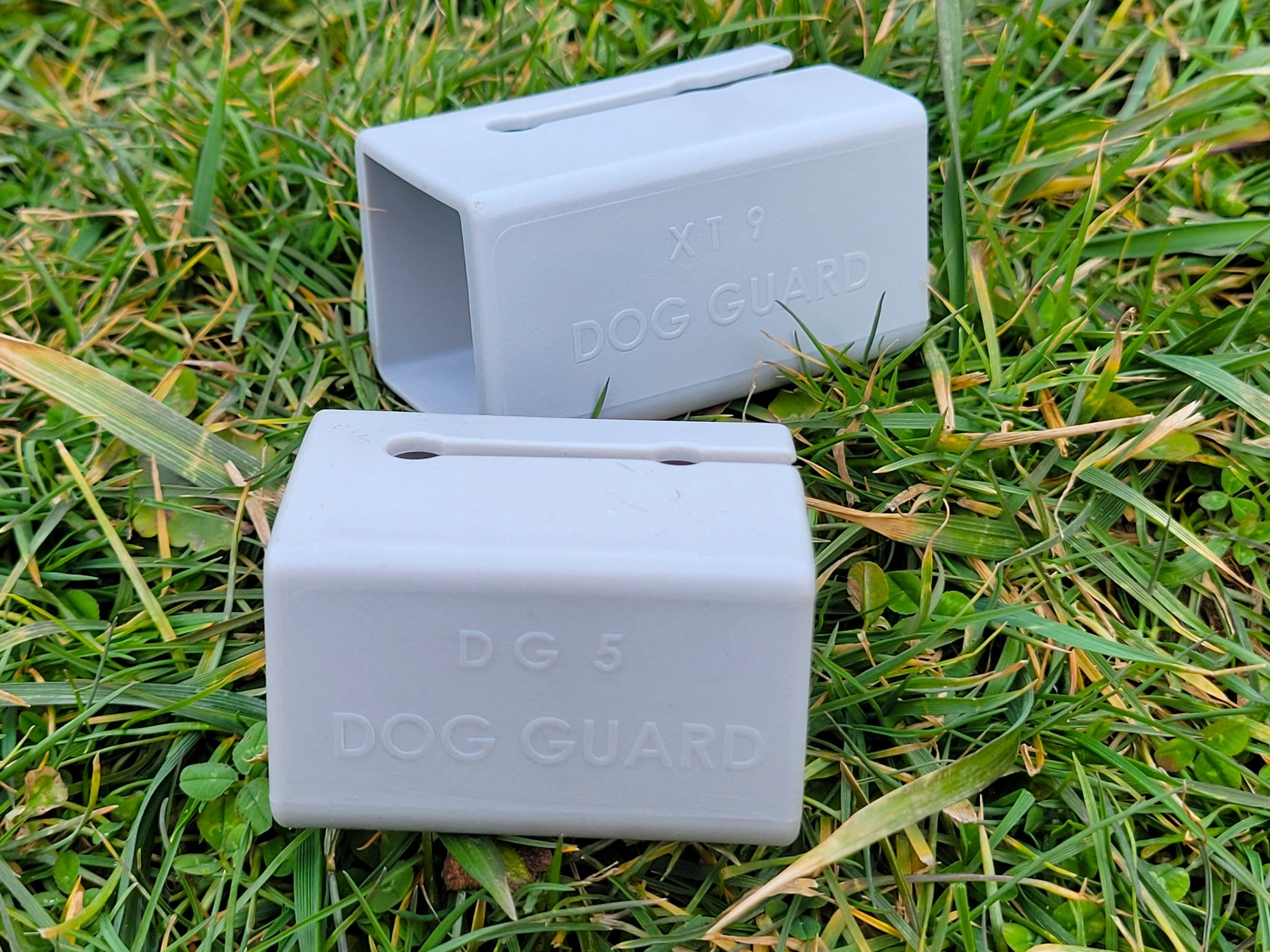 A pair of white plastic dog guard markers with oval openings, placed on green grass. One marker is upright, showing the inscription "XT9 DOG GUARD." The other marker is partially visible in the foreground.