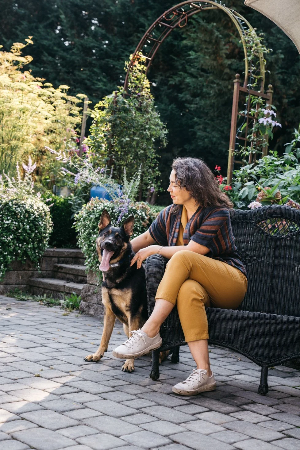 A woman sitting on a black wicker bench in a garden with her German Shepherd dog, both looking content. The garden features lush green plants, flowering bushes, and a decorative metal garden arch in the background.