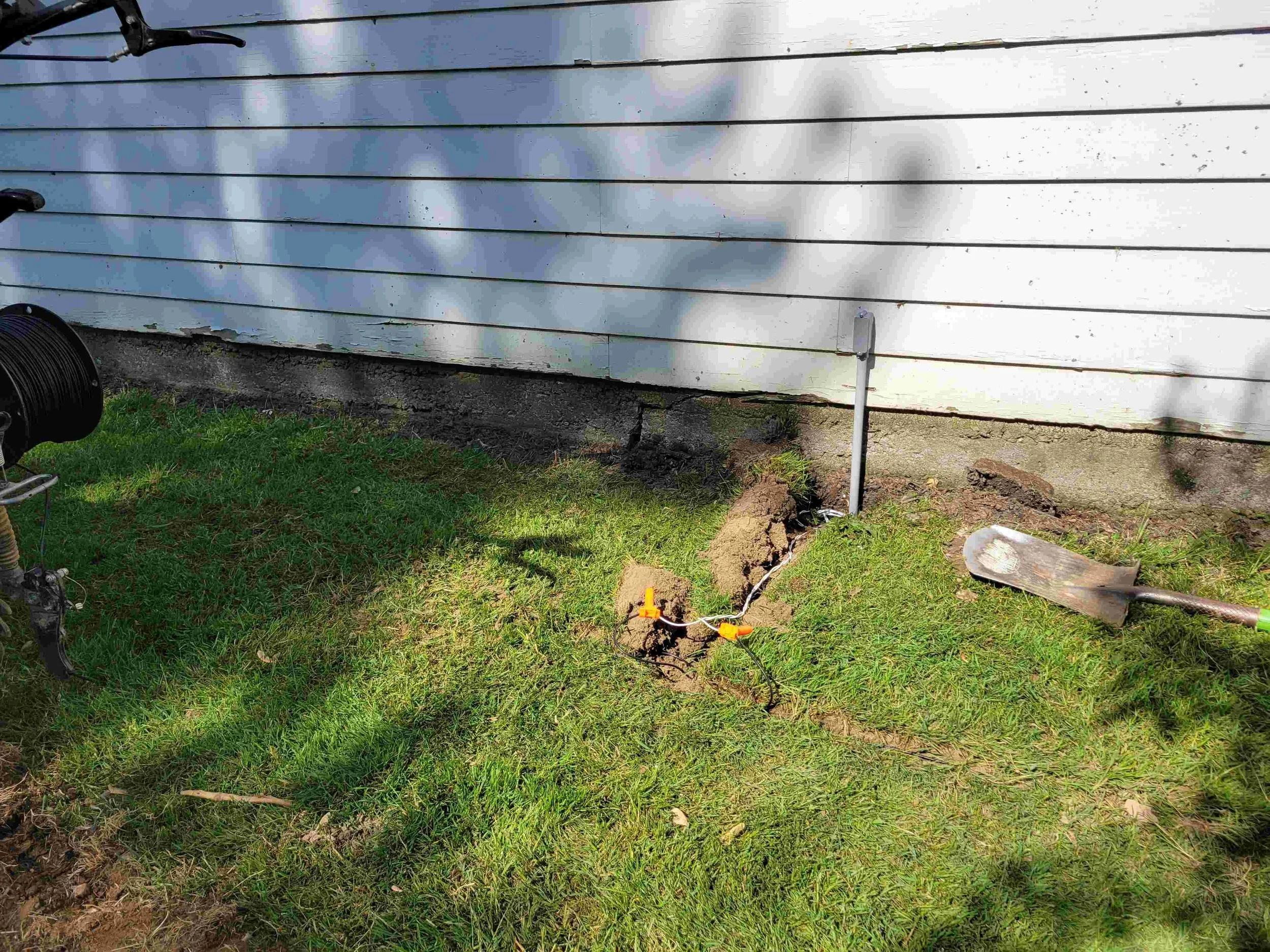 Exterior of a house with white horizontal siding, showing damage at the bottom where the siding is peeling and cracked, with a hole in the foundation. There is a grass lawn in the foreground and shadows of tree branches.