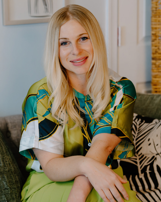 A woman with blonde hair and a bright smile sitting on a couch in a room with light blue walls and framed art. She wears a colorful, patterned shirt with shades of green, blue, white, and black.