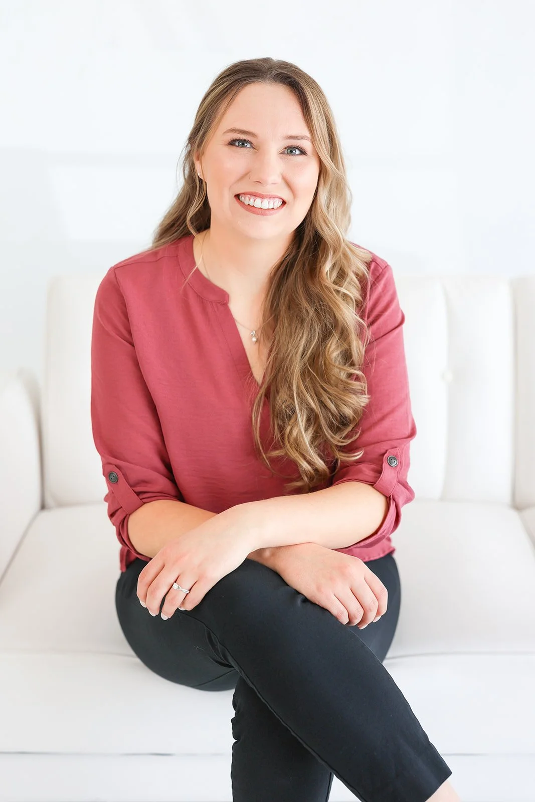 A smiling woman with long wavy hair, wearing a pink blouse and black pants, sitting on a white couch against a plain white wall.