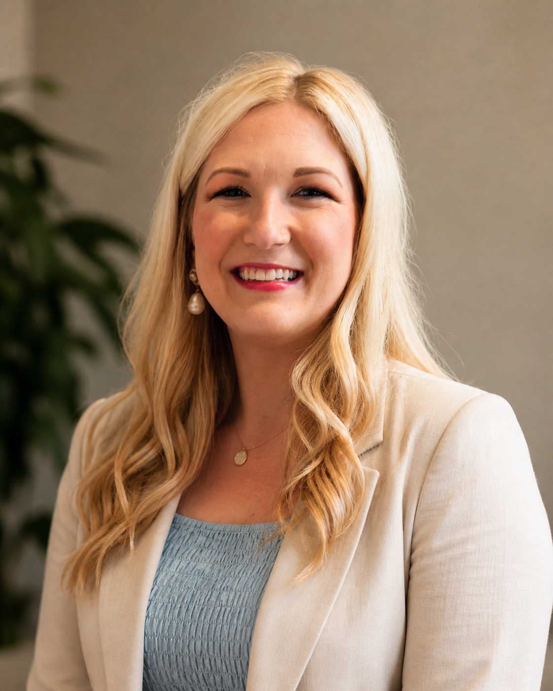 A smiling woman with long blonde hair, wearing a cream blazer, a light blue top, pearl earrings, and a gold necklace, standing indoors with a blurred plant background.