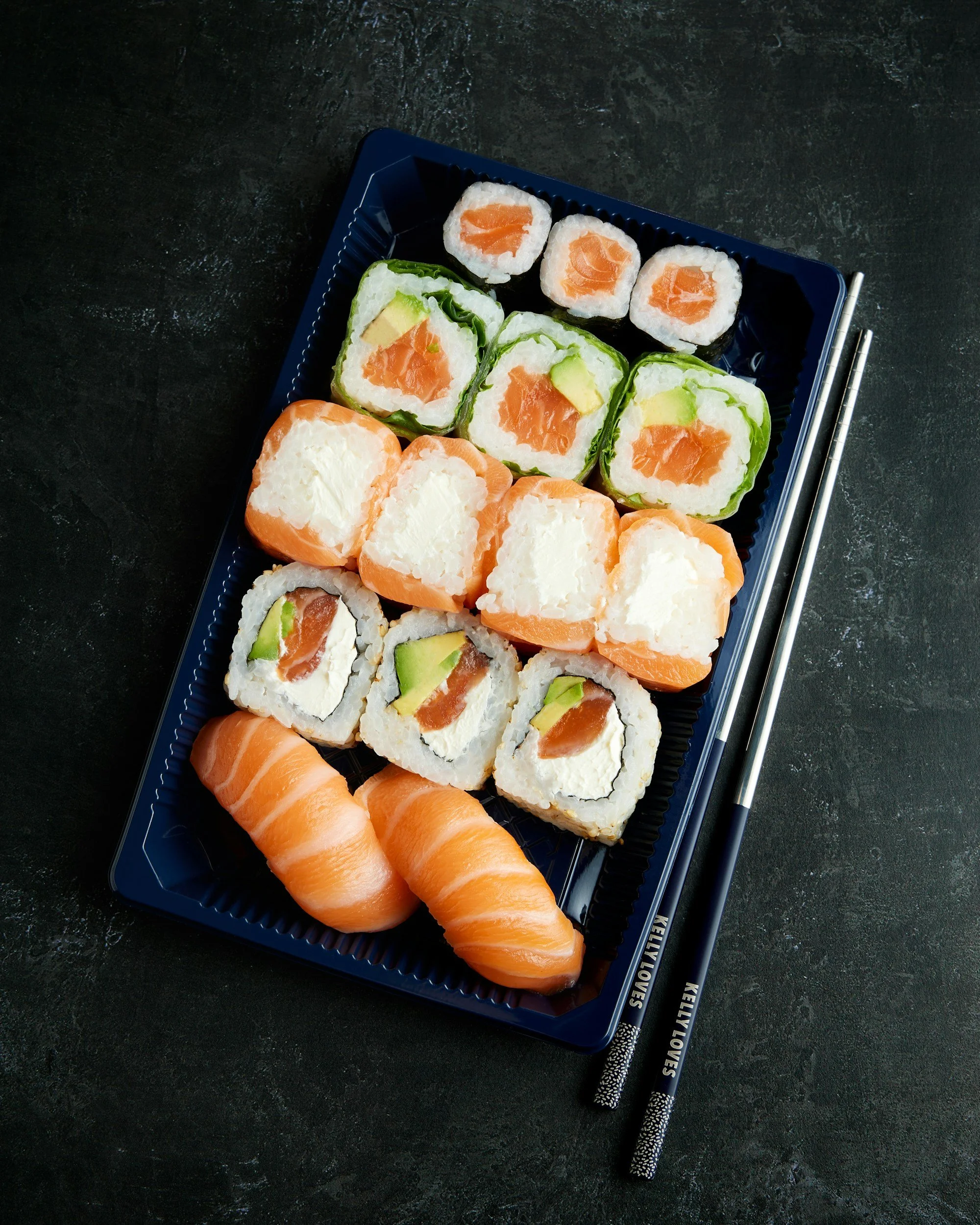 Verschiedene Sushi-Stücke auf einem blauen Teller mit Essstäbchen. Das Bild zeigt Lachs-, Avocado-, und Frischkäsesushi auf einem schwarzen Hintergrund.