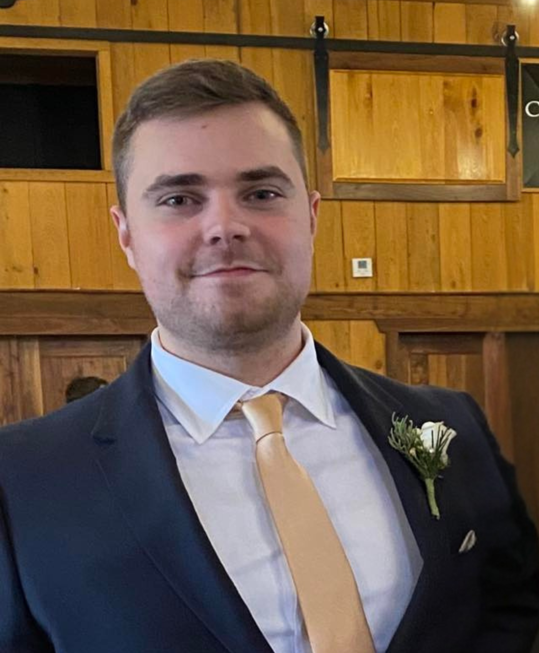A man in formal attire, including a dark suit, white shirt, and cream tie, standing indoors with wooden walls and a railing in the background.