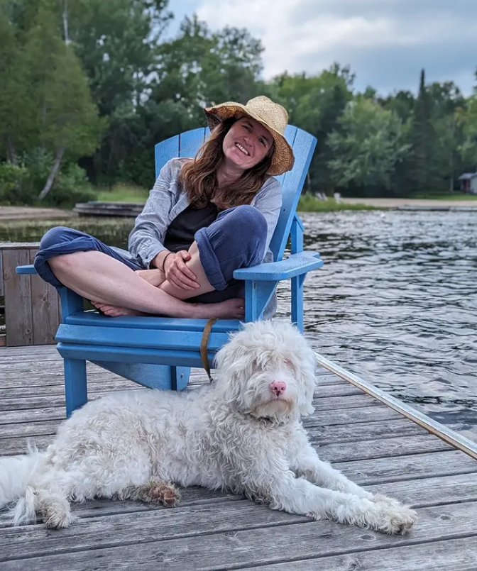 A woman sitting on a bright blue wooden chair on a dock by the water, smiling, wearing a straw hat and casual clothing. A large, fluffy white dog with a pink nose is lying on the dock in front of her, facing the camera. The background features green trees and a cloudy sky.