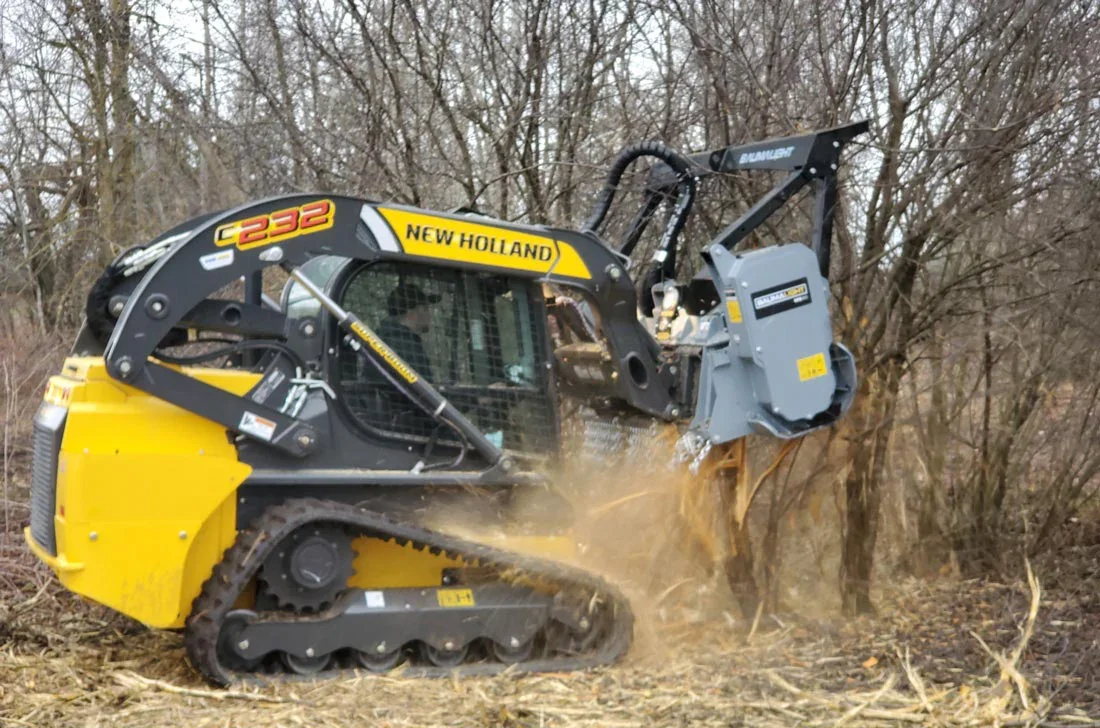 A yellow and gray New Holland C232 compact excavator working in a wooded area, digging into the ground with its bucket.