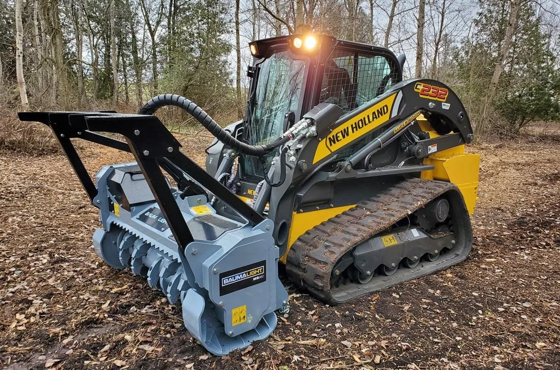 A New Holland compact excavator with a Baumalight mulching attachment working on a dirt and leaf-covered ground in a wooded area.