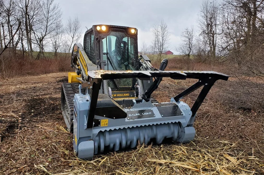 A compact excavator with a mulching attachment on a cleared outdoor area with leafless trees and overcast sky in the background.