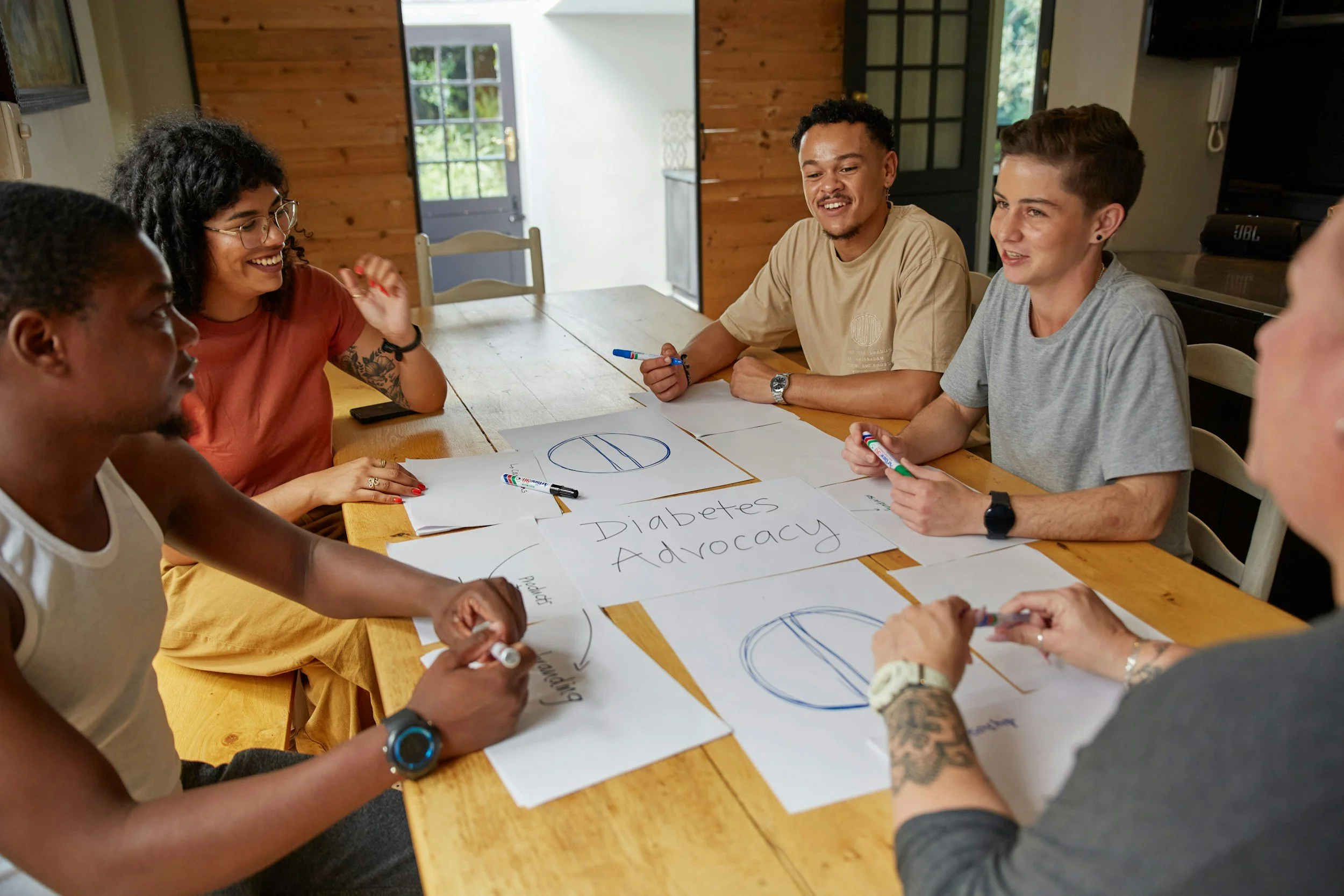 Group of diverse young adults around a wooden table participating in a discussion about diabetes advocacy, with posters displaying the words 'Diabetes Advocacy' and related diagrams visible on the table.
