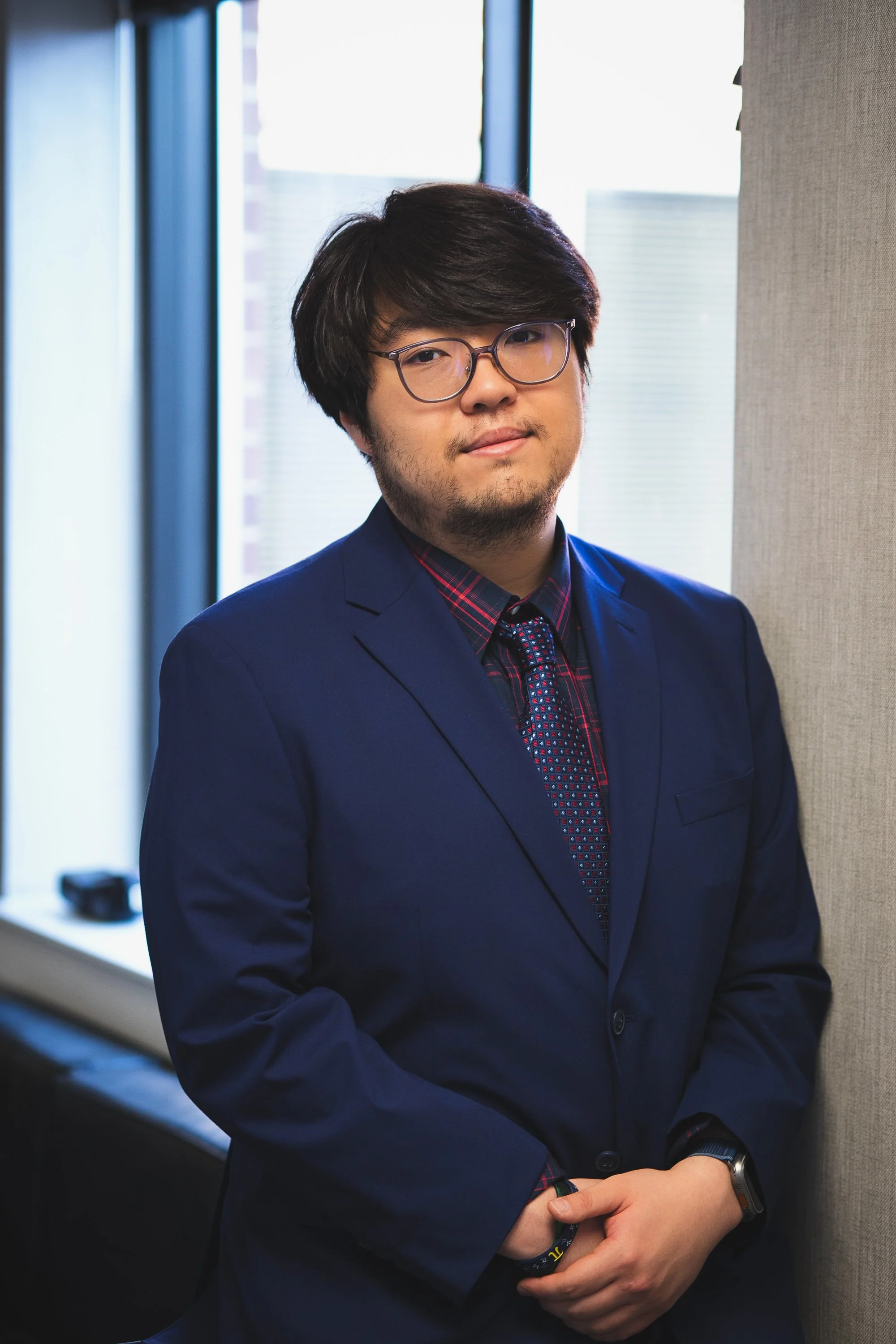A young man with dark hair, glasses, and a light beard dressed in a navy suit with a red checkered shirt and tie, standing indoors near a wall with a window behind him, with his arms crossed and wearing a wristwatch and a wristband.
