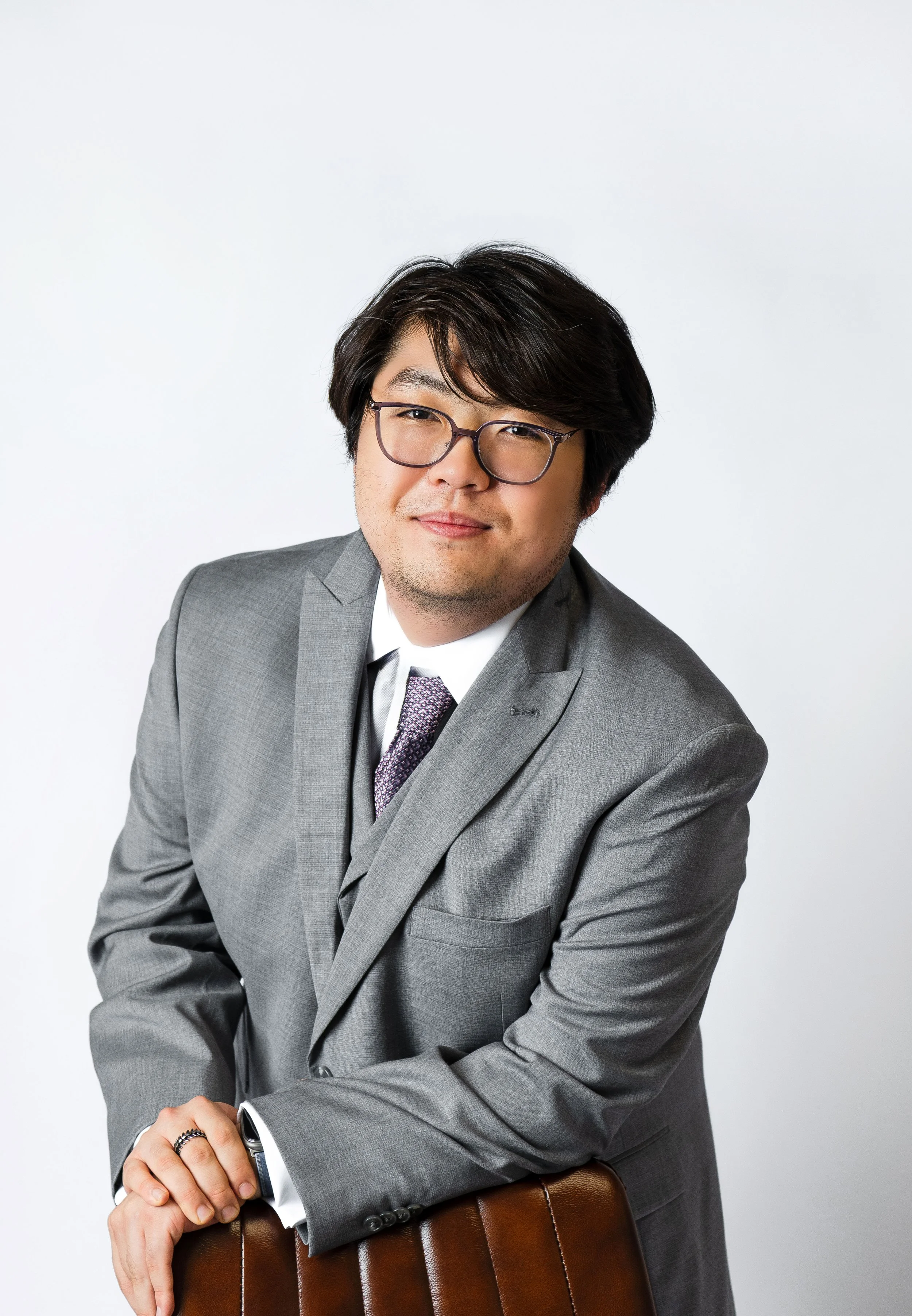Portrait of a man in a gray suit and glasses, sitting on a brown leather chair, smiling slightly against a plain white background.