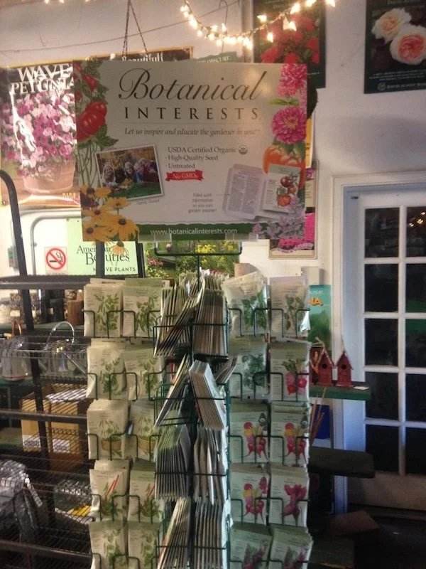 Display stand with packets of botanical interest seeds at a garden center. Sign above promotes inspiring gardens with organic, high-quality seed and plant information.