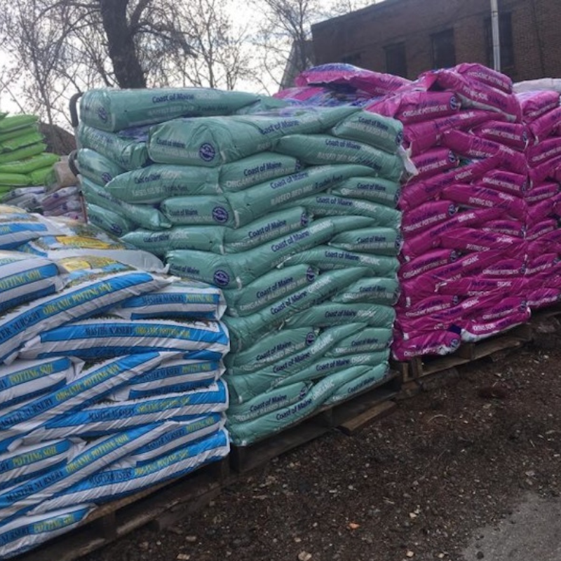 Stacks of colorful bags of soil or fertilizer on pallets outside, including green, pink, and blue bags, with a building and trees in the background.