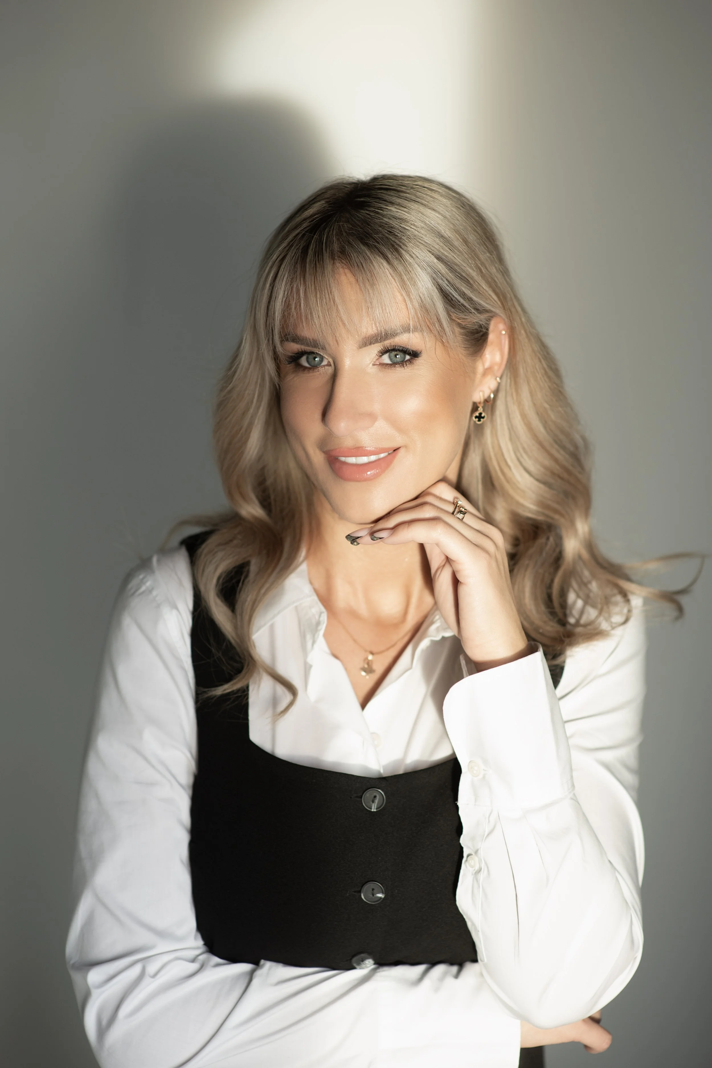 A woman with blonde hair and blue eyes, smiling, wearing a white blouse under a black dress, with earrings and a ring, posing with her hand on her chin against a gray background.