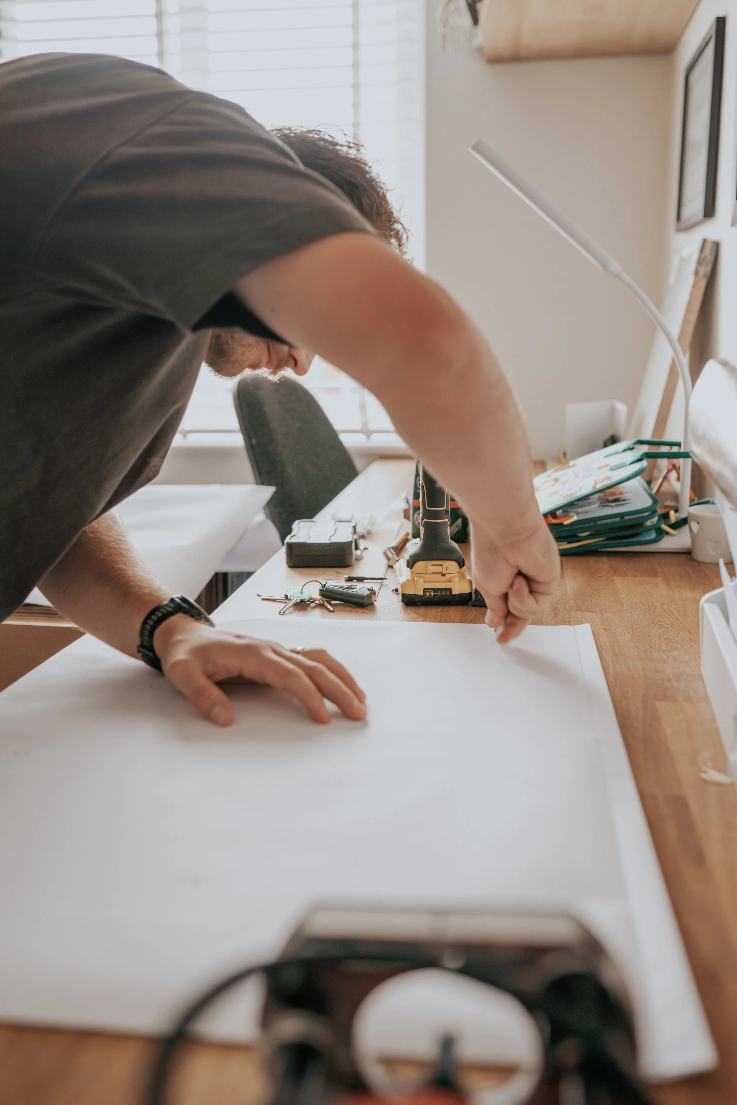A person working on a large sheet of paper on a wooden desk with tools and stationery around, in a room with windows and picture frames on the wall.