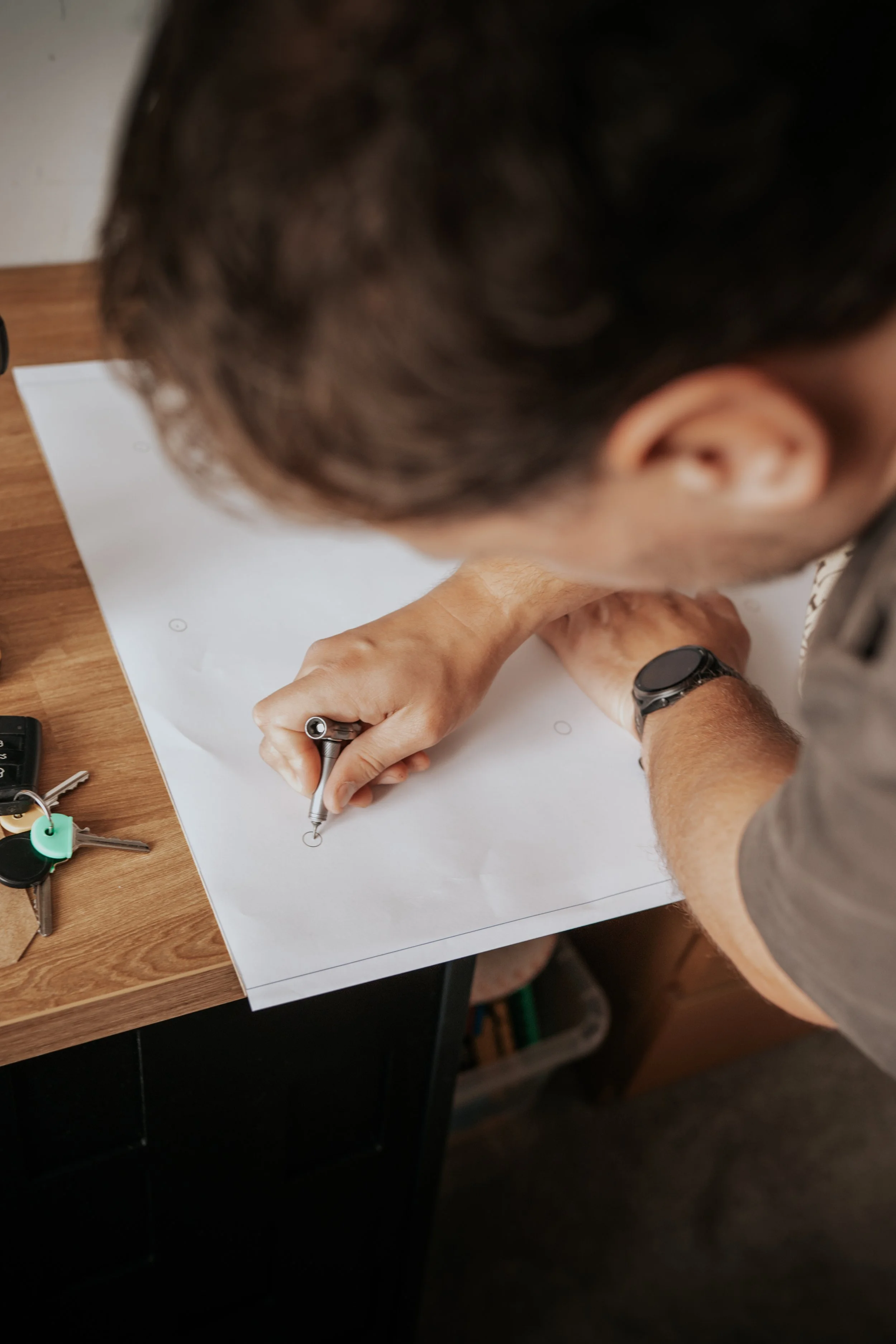 A person with dark hair, wearing a gray shirt and a black watch, is drawing on a large sheet of white paper on a wooden desk using a black pen. There are keys and a black object on the desk.